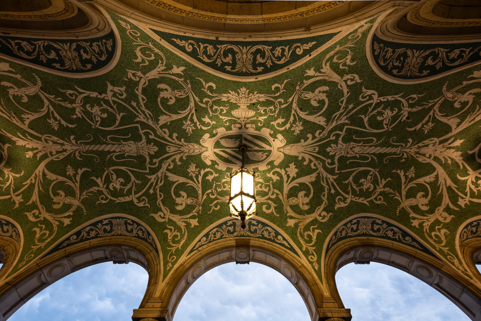 Ornate lamps and a scrolled-mural ceiling of the arched, second-floor portico of the Memorial Union at the University of Wisconsin–Madison are pictured as dusk falls to nighttime.